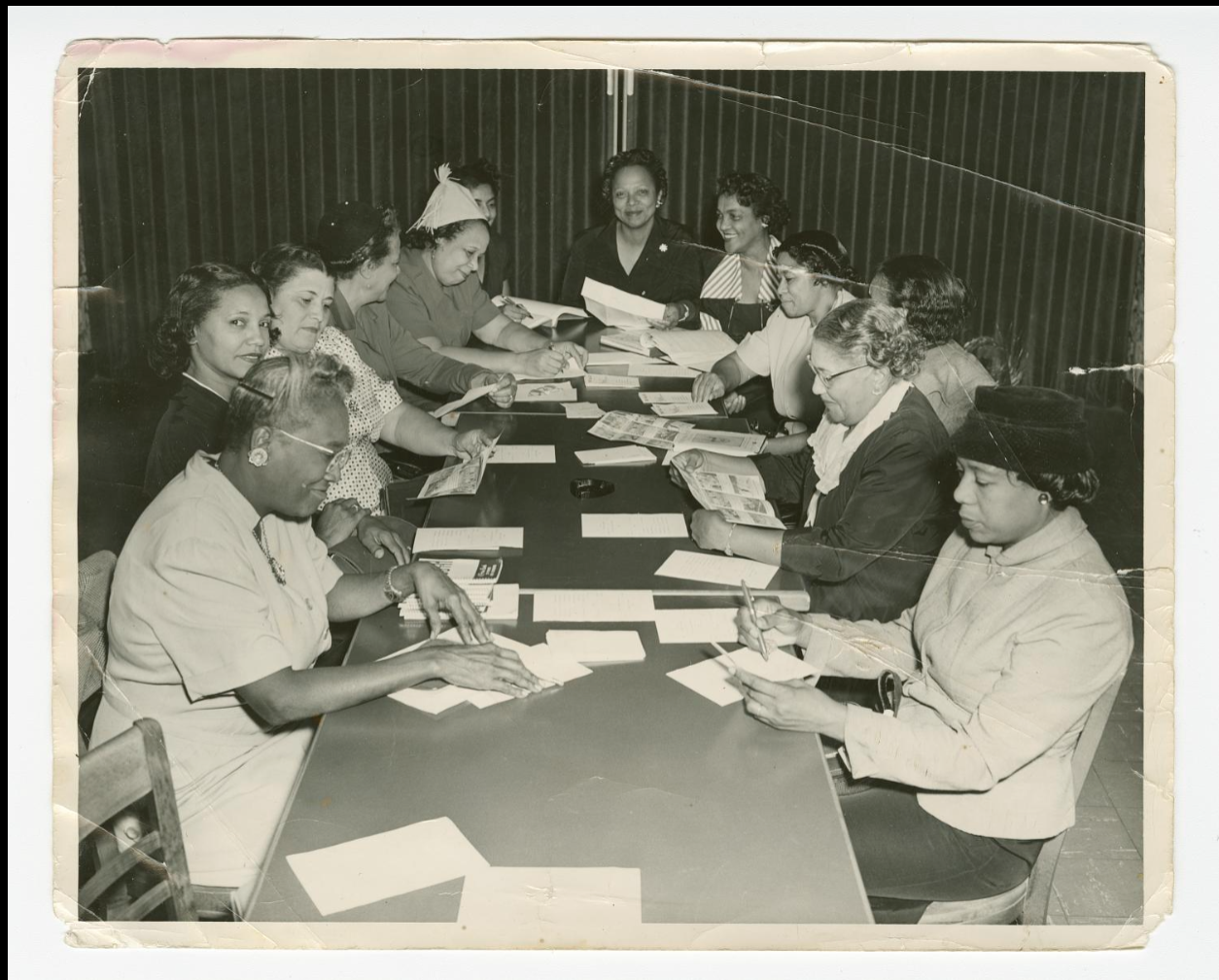 Archival photograph of a group of women seated around a long table reviewing papers and documents together, part of the Princetta R. Newman Collection of Family Photographs